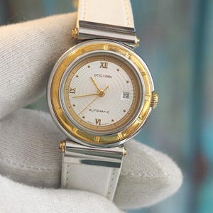 May include: A silver and gold two-tone wristwatch with a white leather band. The watch face is white with Roman numeral hour markers and a date window at the 3 o'clock position. The watch face also has the text "OTTO KERN AUTOMATIC".