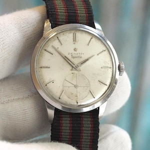 May include: A vintage Zenith Sporto watch with a silver face and a black, green, and red striped nylon strap. The watch face has silver hands and markers. A classic timepiece.