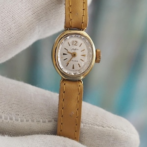 Op de afbeelding: Een gouden polshorloge met een witte wijzerplaat en zwarte wijzers. Het horloge heeft een bruin leren bandje en een kleine secondewijzer.