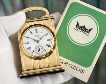 May include: A gold-colored Smiths Astral mantel clock with a white face and black hands. The clock is being held in a white glove. The clock is accompanied by a green card with the text "Smiths Astral" and "Our Clocks".