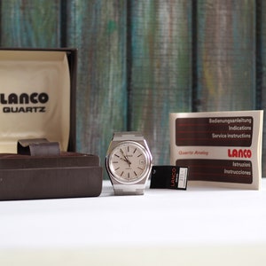May include: A silver Lanco Quartz analog watch with a white face and black hands. The watch is in its original box with instructions in multiple languages.