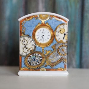 May include: A white porcelain clock with a blue and gold design featuring multiple clock faces. The clock has a Roman numeral clock face, a smaller clock face with Arabic numerals, and a clock face with a compass design.