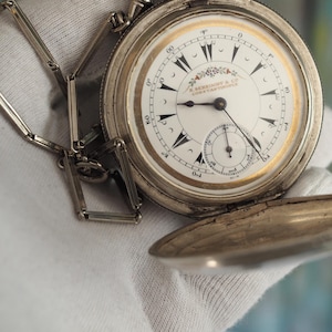 May include: A silver pocket watch with a white face and black Roman numeral hour markers. The watch has a small second hand dial at the bottom of the face. The watch is open and the chain is attached.