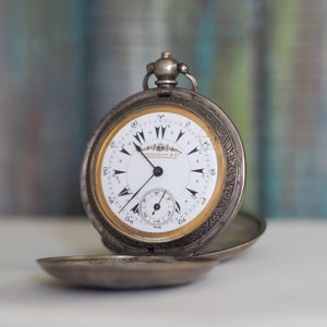 May include: A silver pocket watch with an ornate design and a white face with black Roman numeral markings. The watch is open and the face is visible. The watch is on a white surface.