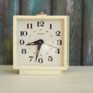 Pode incluir: Relógio despertador quadrado branco com números e ponteiros pretos. O mostrador do relógio diz "SEVANI" e tem um ponteiro das horas preto apontando para o 8 e um ponteiro dos minutos preto apontando para o 12.