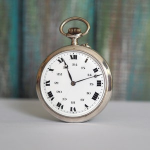 Könnte beinhalten: Eine silberne Taschenuhr mit römischen Ziffern auf einem weißen Zifferblatt. Die Uhr hat eine silberne Kette, die oben befestigt ist.