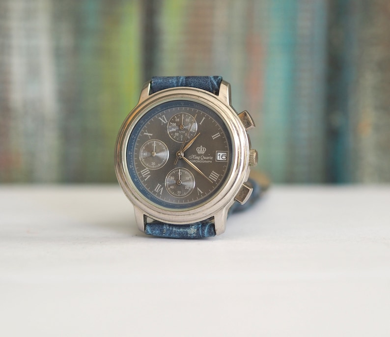 Pu&ograve; includere: Un orologio da polso cronografo argentato con quadrante blu e cinturino in pelle blu. Il quadrante dell'orologio ha indici orari in numeri romani e una finestrella per la data alle 4. L'orologio &egrave; etichettato "King Quartz Chronograph".