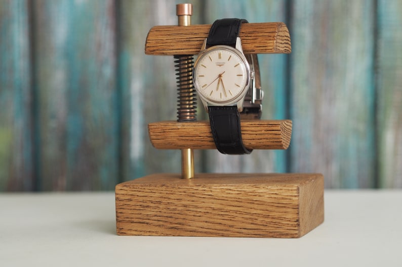 May include: A wooden watch stand with a silver watch with a black leather band. The stand has a brass screw and a wooden base.