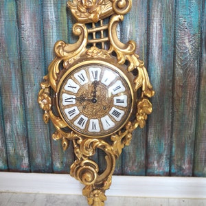 Puede incluir: Reloj de pared dorado y ornamentado con un diseño detallado y en espiral. La esfera del reloj tiene números romanos y filigranas doradas decorativas. El reloj mide aproximadamente 30 cm de alto.