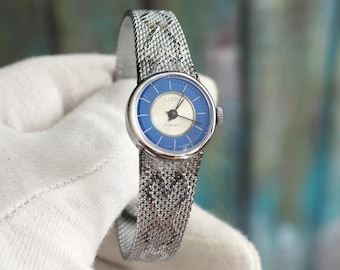 May include: A vintage wristwatch with a silver-tone case and a blue and white dial. The watch features a silver mesh band. The dial has the word "Luch" printed on it. The watch is held by a gloved hand.