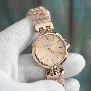 May include: A rose gold wristwatch with a light pink face and a matching beaded strap. The watch face has the text "Pierre Chaubert". The strap is adorned with small pink beads and clear crystals.