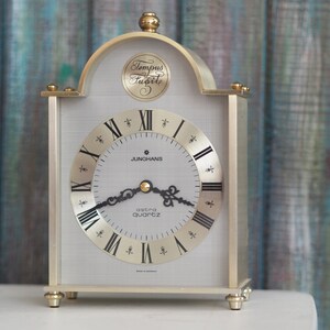 May include: A gold-toned analog clock with Roman numeral markings on the face. The clock face reads "Junghans Astra Quartz". The clock has a decorative top with the inscription "Tempus Fugit".