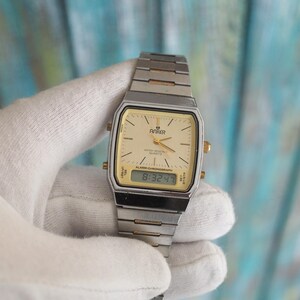 May include: A silver and gold tone analogue and digital wristwatch with a white face and black digital display. The watch face has the text "Anker" and "Alarm-Chronograph" on it. The digital display shows the time "8:32:47".
