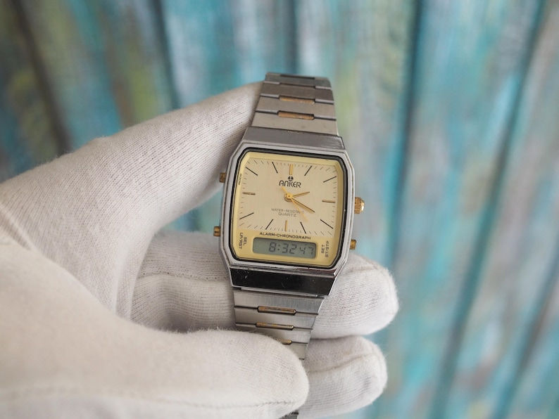 May include: A silver and gold tone analogue and digital wristwatch with a white face and black digital display. The watch face has the text "Anker" and "Alarm-Chronograph" on it. The digital display shows the time "8:32:47".