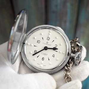May include: A silver pocket watch with a white face and black hands. The watch has a chain attached to it and says "Cortebert 17 Jewels Incabloc" on the face.