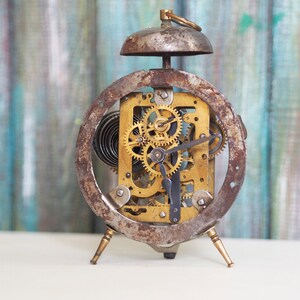 Könnte beinhalten: Ein alter, rostiger Wecker mit freiliegenden Zahnrädern und einer Glocke oben. Der Wecker steht auf einer weißen Oberfläche mit einem blau-grünen gestreiften Hintergrund.