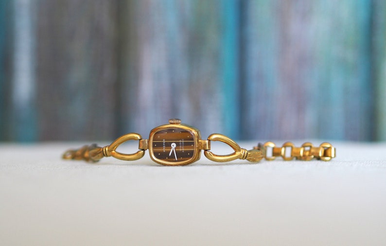 May include: A gold-toned wristwatch with a square face and a brown tiger's eye dial. The watch has a gold-toned metal band with a unique link design.