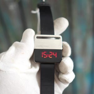 May include: A digital wristwatch with a black band and a rectangular face. The top half of the face is silver, with the text "CHEITEL PARIS" printed on it. The bottom half of the face displays the time in red digital numbers: 15:24.