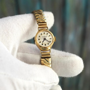 Può includere: Un orologio da polso vintage color oro con quadrante bianco e lancette nere. L'orologio presenta un cinturino dorato con un design a spina di pesce e un piccolo quadrante rotondo. Il quadrante ha piccoli indicatori quadrati e il marchio "Tullem" è visibile.