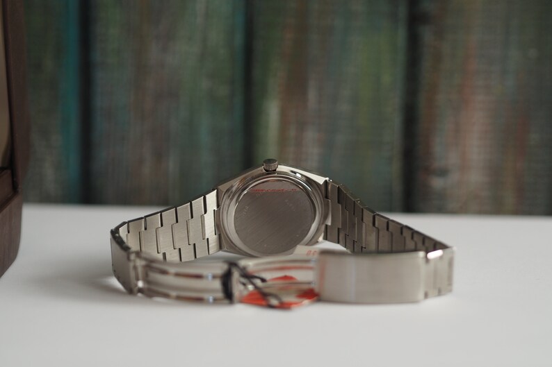 May include: A silver wristwatch with a stainless steel band. The watch has a round face and a silver case. The back of the watch is visible in the image.