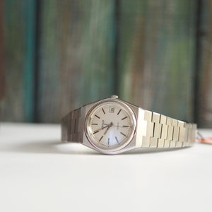 May include: A silver wristwatch with a white face and a silver metal band. The watch has a date window at the 3 o'clock position and the word "Coda" printed on the face.
