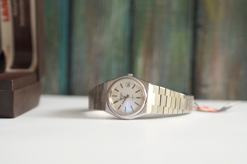May include: A silver wristwatch with a white face and a silver metal band. The watch has a date window at the 3 o'clock position and the word "Coda" printed on the face.