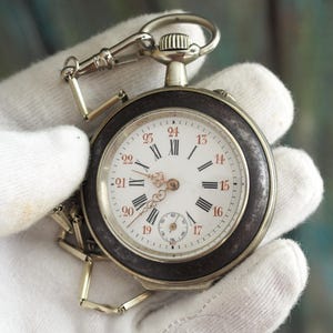May include: Antique pocket watch with a white face, black Roman numerals, and red Arabic numerals. The watch has a silver-colored case and a dark brown bezel. A chain and clasp are attached to the watch.