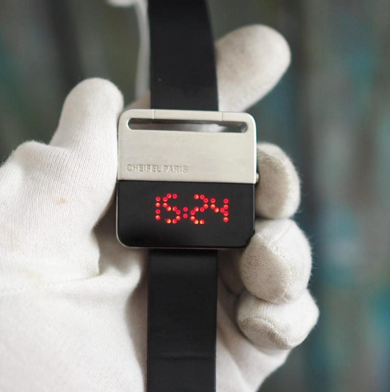 May include: A modern digital watch with a black band and a rectangular face. The watch face displays the time in red LED lights. The watch has a silver-colored clasp and the text "CHEIFEL PARIS".