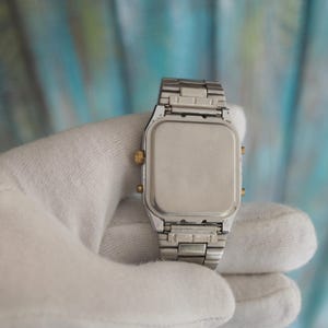 May include: A silver and gold digital watch with a square face and a metal strap. The watch is being held in a white cloth.