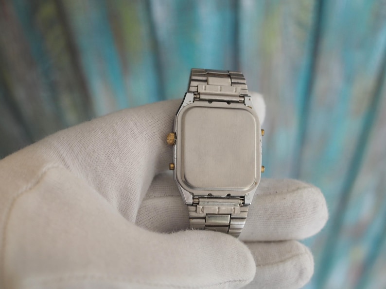 May include: A silver and gold digital watch with a square face and a metal strap. The watch is being held in a white cloth.