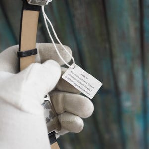 May include: A black wristwatch with a leather band is held by a gloved hand. A white tag attached to the watch displays text, including the words "Made for" and the address of a German company.