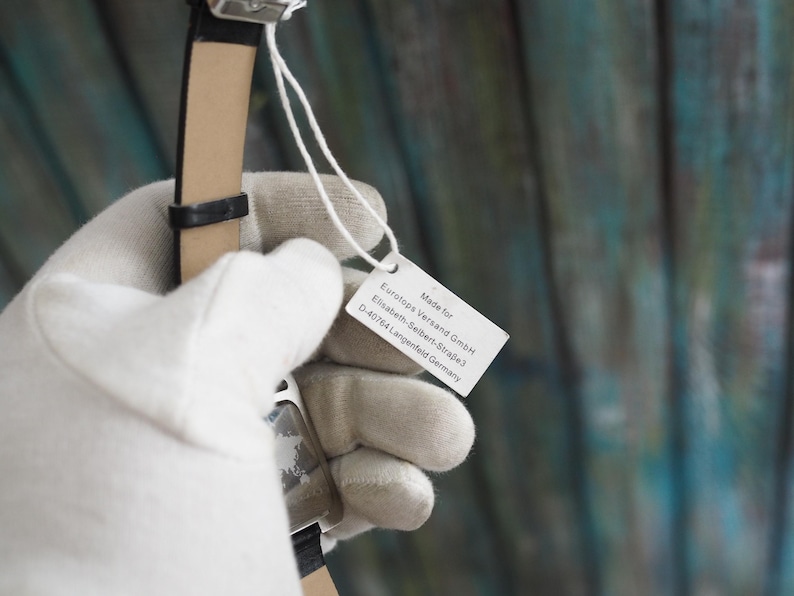 May include: A black wristwatch with a leather band is held by a gloved hand. A white tag attached to the watch displays text, including the words "Made for" and the address of a German company.