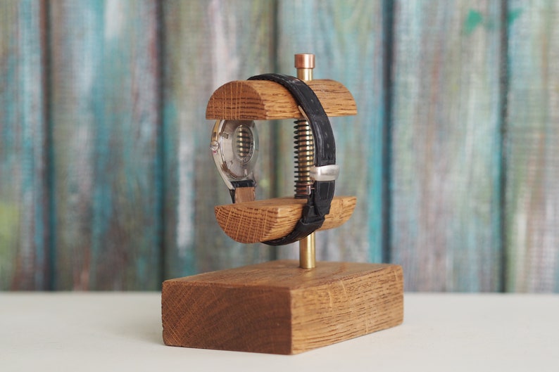 May include: A wooden watch stand with a watch and a black leather strap. The stand is made of oak wood and has a brass screw.