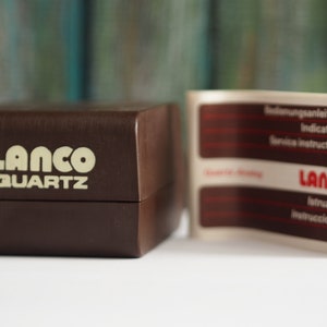 May include: A brown box with the words "LANCO QUARTZ" printed in white on the front. A white paper with red text is attached to the box. The paper has the words "Bedienungsanleitung", "Indications", "Service Instructions", "LANCO", "Istruzioni", and "Instrucciones" printed on it.