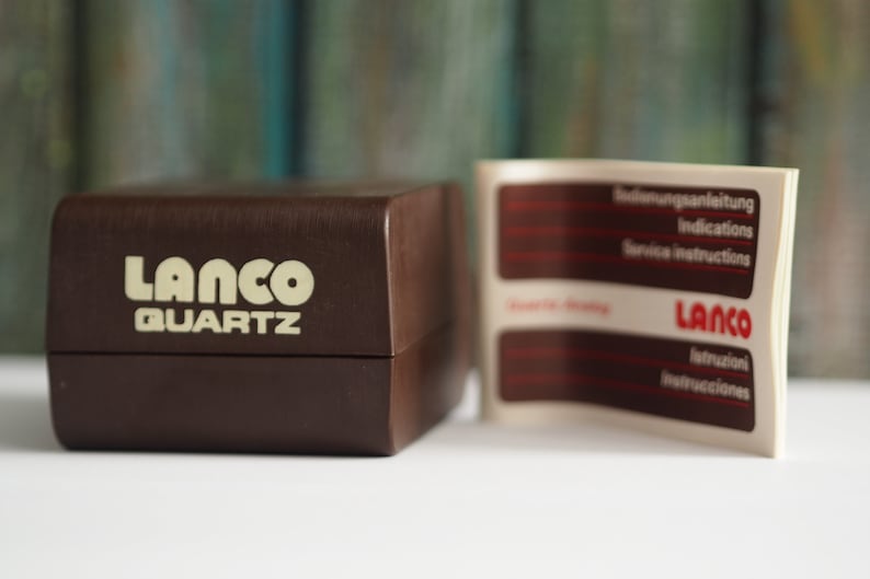 May include: A brown box with the words "LANCO QUARTZ" printed in white on the front. A white paper with red text is attached to the box. The paper has the words "Bedienungsanleitung", "Indications", "Service Instructions", "LANCO", "Istruzioni", and "Instrucciones" printed on it.