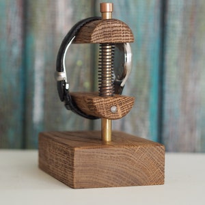 May include: A wooden watch stand with a brass screw and a black leather watch band. The stand is made of oak wood and has a rectangular base.