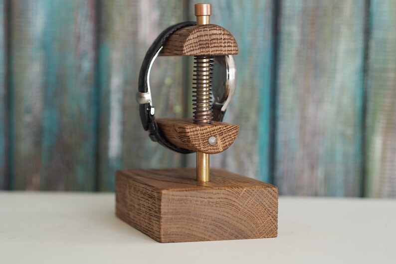 May include: A wooden watch stand with a brass screw and a black leather watch band. The stand is made of oak wood and has a rectangular base.