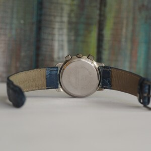 Pu&ograve; includere: Un orologio da polso argentato con un cinturino in pelle blu. L'orologio ha una funzione cronografo e una cassa in acciaio inossidabile.