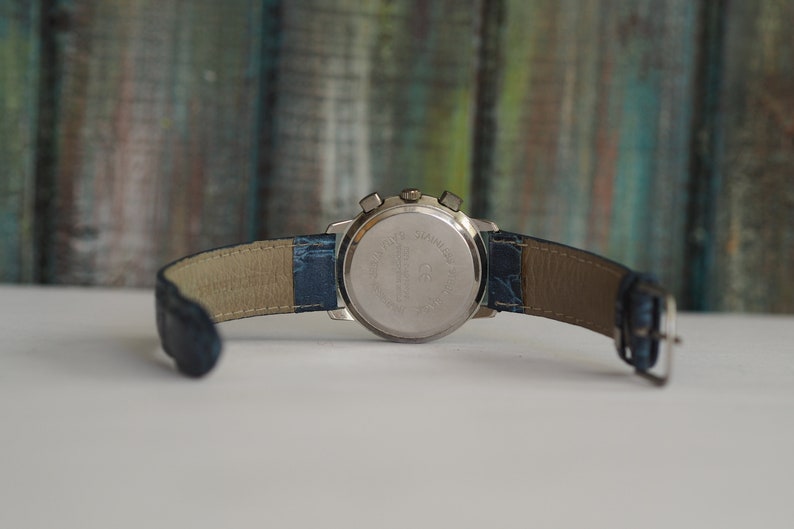 Pu&ograve; includere: Un orologio da polso argentato con un cinturino in pelle blu. L'orologio ha una funzione cronografo e una cassa in acciaio inossidabile.