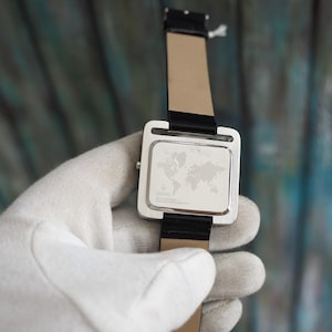 May include: A silver wristwatch with a black leather band. The watch face is square with a world map design. The watch is held by a gloved hand.