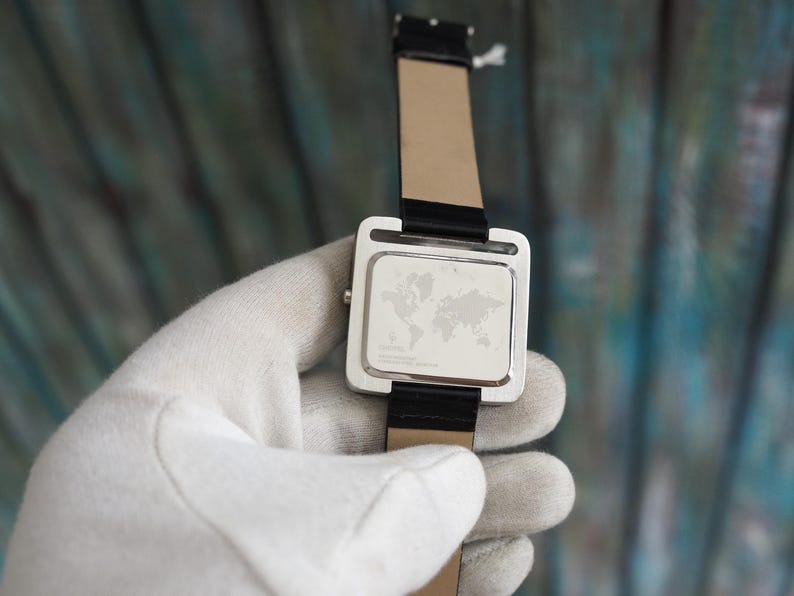 May include: A silver wristwatch with a black leather band. The watch face is square with a world map design. The watch is held by a gloved hand.