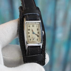 May include: A vintage wristwatch with a rectangular silver case and a cream face. The watch has black numerals and hands, and a small seconds dial. It's attached to a wide black leather cuff-style strap.