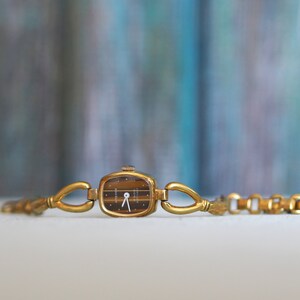 May include: A gold-toned wristwatch with a square face and a black dial. The watch has a delicate chain link bracelet.