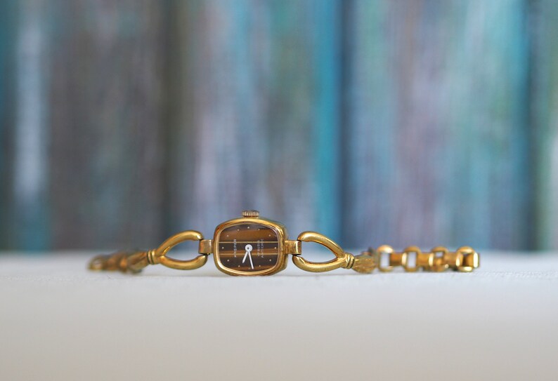 May include: A gold-toned wristwatch with a square face and a black dial. The watch has a delicate chain link bracelet.