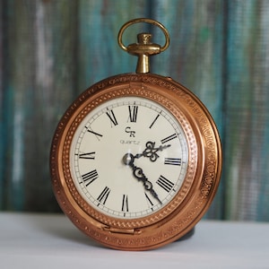 Puede incluir: Un reloj de bolsillo de color cobre con marcas de números romanos en la esfera. La esfera del reloj es blanca con manecillas negras y el texto "CR quartz" en la parte superior.