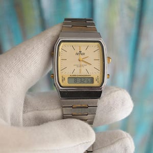 May include: A silver and gold tone analogue and digital wristwatch with a white face and black digital display. The watch face has the text "Anker Water Resistant Quartz" and "Alarm-Chronograph". The digital display shows the time "8:32:53".