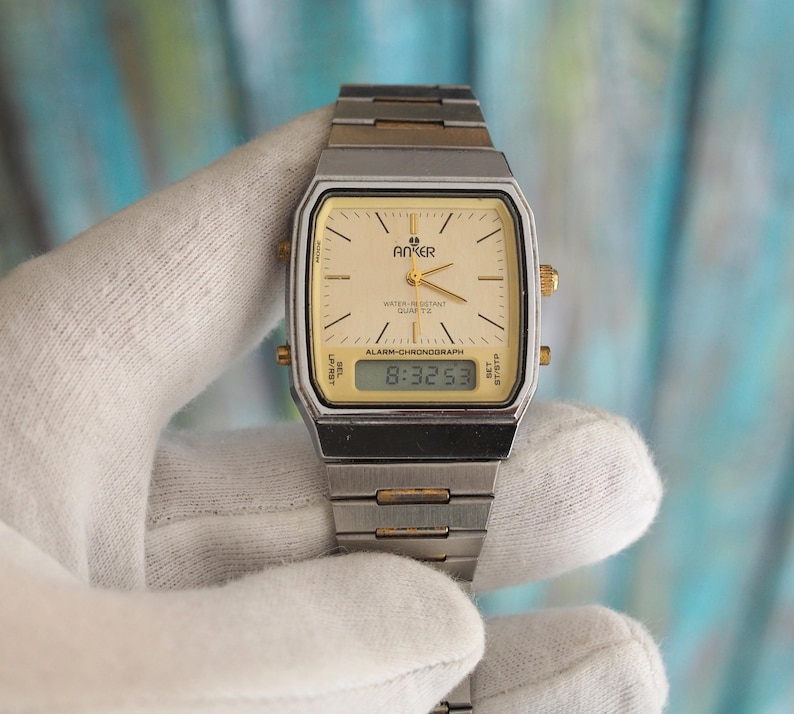 May include: A silver and gold tone analogue and digital wristwatch with a white face and black digital display. The watch face has the text "Anker Water Resistant Quartz" and "Alarm-Chronograph". The digital display shows the time "8:32:53".