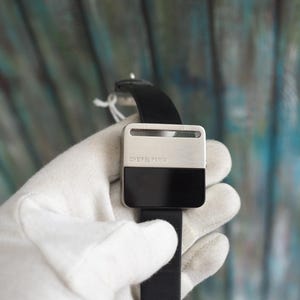 May include: A square-faced watch with a black leather band. The watch face is divided into two sections: a silver top and a black bottom. The text "CHEIFEL PARIS" is printed on the silver section.
