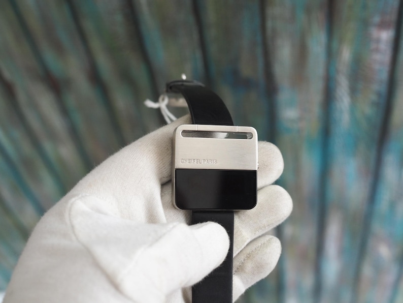 May include: A square-faced watch with a black leather band. The watch face is divided into two sections: a silver top and a black bottom. The text "CHEIFEL PARIS" is printed on the silver section.