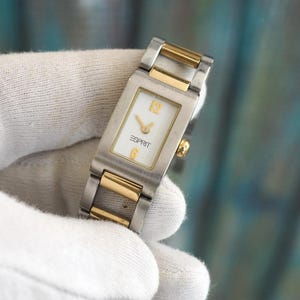 Puede incluir: Un reloj de pulsera de dos tonos, plateado y dorado, con una esfera blanca y agujas negras. La esfera del reloj tiene la palabra "ESPRIT" impresa.